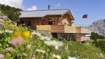 Vue extérieure du restaurant de montagne Sareis avec des fleurs au premier plan et des montagnes en arrière-plan Vue extérieure du restaurant de montagne Sareis avec des fleurs au premier plan et des montagnes en arrière-plan