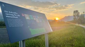 Panneau d'information sur le champ alimentaire de Vaduz avec vue sur les Alpes au coucher du soleil - un lieu d'apprentissage sur l'alimentation durable au Liechtenstein.