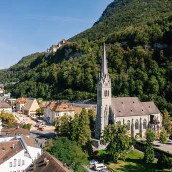 Vue aérienne de la cathédrale Saint-Florin à Vaduz avec le château de Vaduz en arrière-plan, par une journée ensoleillée.