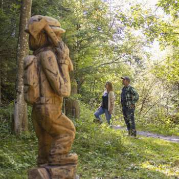 Deux randonneurs découvrent un personnage sculpté sur le « sentier des légendes walsers » au milieu d'une forêt baignée de lumière.