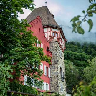 Maison rouge historique à Vaduz, nichée dans une nature verdoyante avec vue sur les montagnes environnantes et un ciel légèrement nuageux.