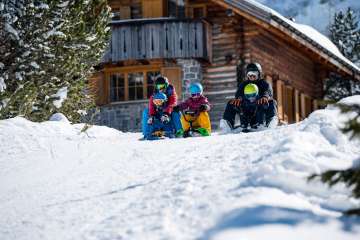 Deux adultes et trois enfants assis sur des luges entament ensemble une descente devant un chalet de montagne enneigé à Malbun.