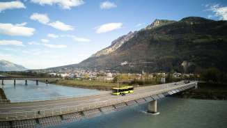 Un bus jaune LIEmobil traverse le pont sur le Rhin près de Vaduz avec un panorama de montagnes en arrière-plan - Découvrir le Liechtenstein