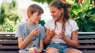 Deux enfants sont assis sur un banc dans un parc du Liechtenstein et mangent une glace - des visages souriants, des fleurs et une ambiance estivale dans la verdure.