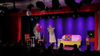 Scène de cabaret au K-BUM au Liechtenstein - scène colorée avec canapé, duo d'acteurs et public dans la salle.