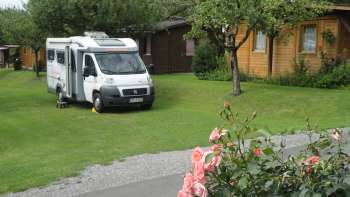 Aire de camping-car sur le camping Mittagsspitze avec une pelouse verte et des cabanes en bois entourées.