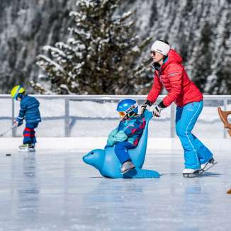 Une mère pousse son enfant sur un pingouin auxiliaire de patinage sur la patinoire de Malbun, avec en arrière-plan un enfant avec une crosse de hockey sur glace.