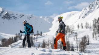 Deux personnes en randonnée hivernale à travers le paysage enneigé de Malbun avec les montagnes enneigées en arrière-plan.