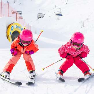 Deux enfants avec des casques roses font du ski en riant sur la piste de Malbun par un temps hivernal ensoleillé.