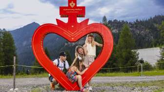Famille posant au cadre rouge du cœur le long du parcours du cœur alpin à Malbun, Liechtenstein, entourée de nature alpine et de montagnes.