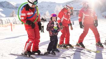 Trois moniteurs de ski en combinaison rouge et un enfant à ski dans la neige, avec des montagnes enneigées et le soleil en arrière-plan.
