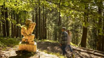 Des randonneurs passent devant un personnage sculpté avec des jumelles au « sentier des légendes walsers »